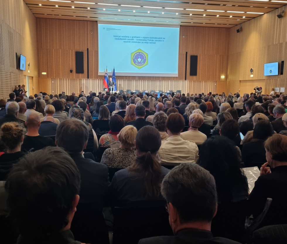 Polna dvorana ljudi. Na odru stoji moški in govori. Zadaj veliko platno, kjer se predvaja predstavitev.
