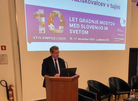 Minister Papič za govorniškim pultom, pred projekcijskim platnom z napisom: Dobrodošli na 10. simpoziju slovenskih raziskovalcev v tujini – 10 let gradnje mostov med Slovenijo in svetom, VTIS simpozij 2025.