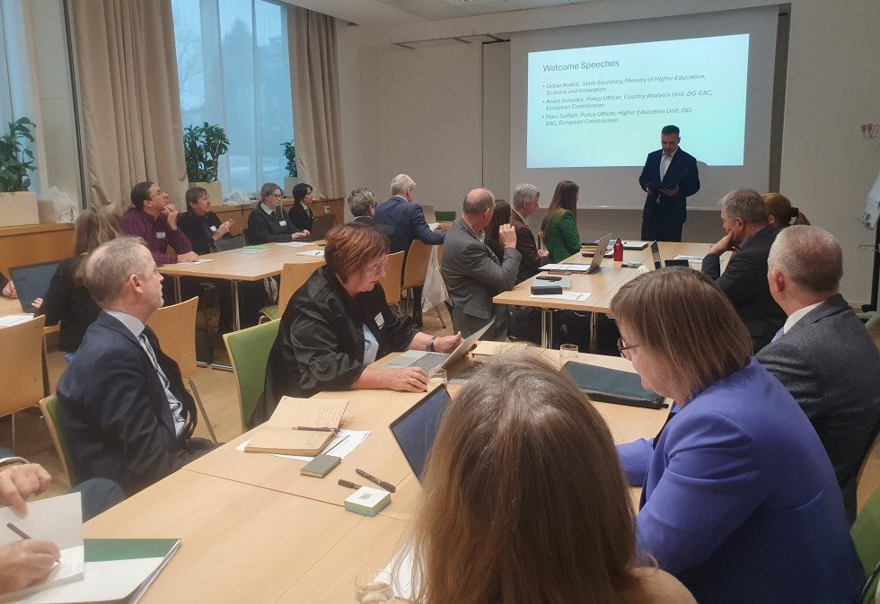 Udeleženci v sejni sobi spremljajo državnega sekretarja Kodriča, ki pred projekcijskim platnom podaja uvodni nagovor.