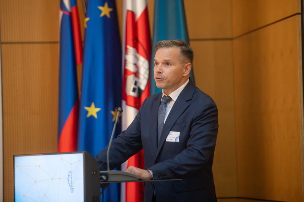 Moški stoji za govornici in ima govor. Zadaj zastave.