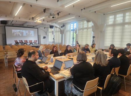 Udeleženci mednarodnega posveta sedijo za veliko mizo z računalniki in dokumenti v dvorani Centra Rog. V ozadju je konferenjski oder s projekcijskim platnom.