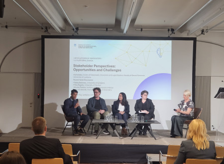 Udeleženec govori v mikrofon med okroglo mizo na konferenčnem odru, ostali panelisti poslušajo razpravo.