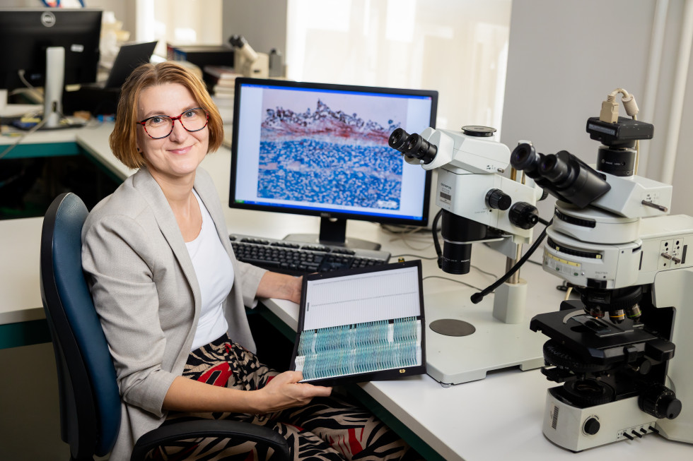 Dr. Jožica Gričar v laboratoriju sedi ob mikroskopu in računalniku ter drži mapo z mikroskopskimi preparati.