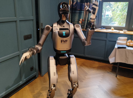 Humanoidni robot stoji v prostoru dogodka in z dvignjenimi rokami pozdravlja udeležence.