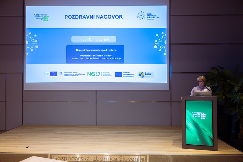 Govornica stoji za govorniškim pultom in nagovarja udeležence dogodka v dvorani Gospodarske zbornice Slovenije.