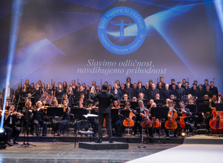  Orkester in zbor pod vodstvom dirigenta na slovesnosti podelitve Zoisovih nagrad. Na osvetljenem odru so glasbeniki in pevci, v ozadju pa je projekcija z napisom "Slavimo odličnost, navdihujemo prihodnost" ter logotip dogodka.
