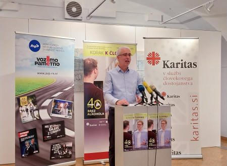 Moški stoji za govorniškim pultom. Za njim roll-upi z vsebino: Korak k človeku, 40 dni brez alkohola, vozimo pametno in Karitas.
