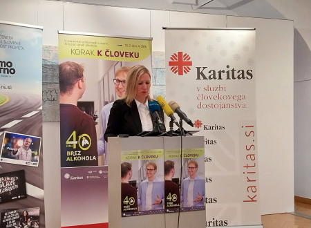 Ženska stoji za govorniškim pultom. Za njo roll-upi: Korak k človeku, 40 dni brez alkohola in Karitas.
