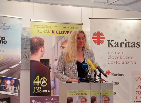 Svetlolasa ženska stoji za govorniškim pultom. Za njo roll-upa z vsebino: Korak k človeku, 40 dni brez alkohola in Karitas.