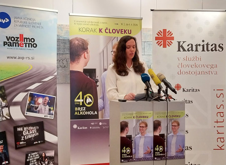 Temnolasa ženska stoji za govorniškim pultom. Za njo roll-upi: Korak k človeku, 40 dni brez alkohola, vozimo pametno in Karitas.