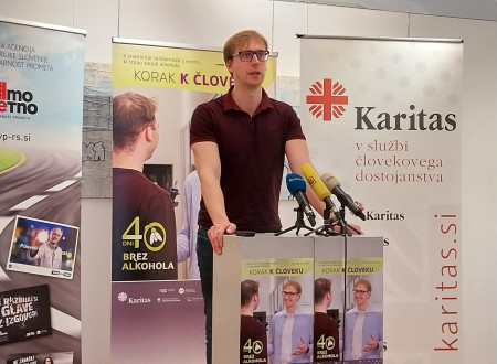 Mlad moški stoji za govorniškim pultom. Za njim roll-upa: Korak k človeku, 40 dni brez alkohola in Karitas.