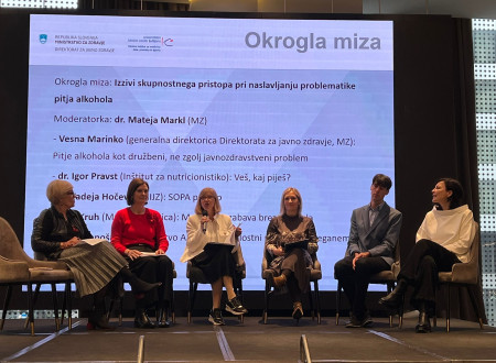 Okrogla miza o skupnostnem pristopu. Nastopajoči sedijo na odru. Za njimi veliko platno s predstavitvijo.