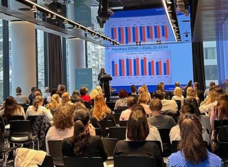 Polna dvorana udeležencev, ki poslušajo predavateljico. Na steni veliko platno s podatki raziskave ESPAD.