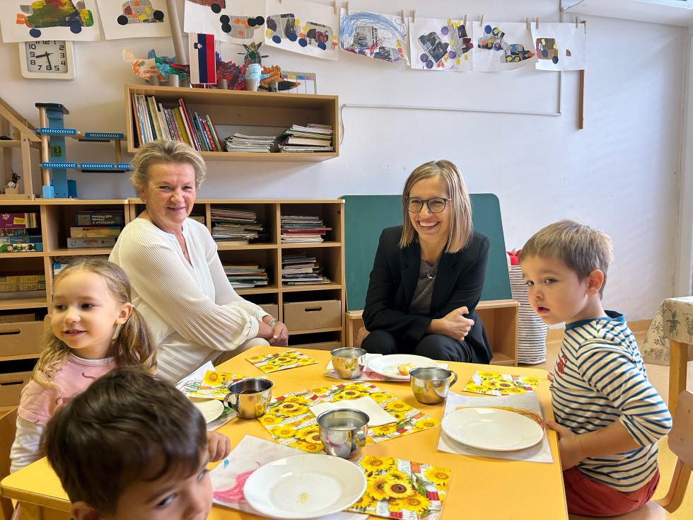 Ministrica za zdravje v vrtcu sedi za nizko mizo s tremi otroki in vzgojiteljico. Pred seboj imajo krožnike, očitno so zajtrk že pojedli.