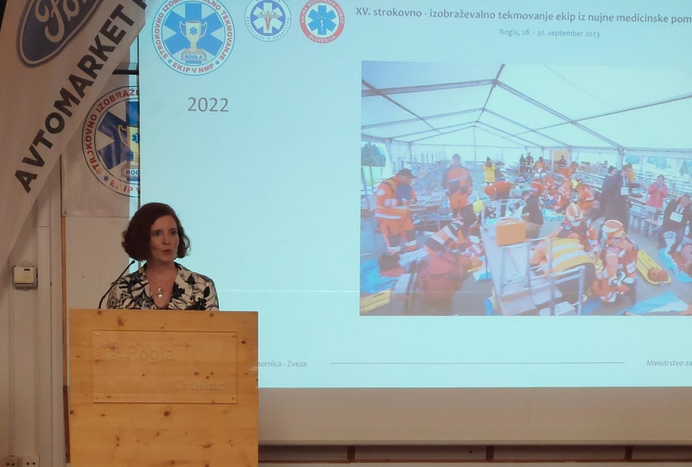 Državna sekretarka Azra Herceg stoji za govorniškim odrom, za njo je projecirana fotografija skupine reševalcev.