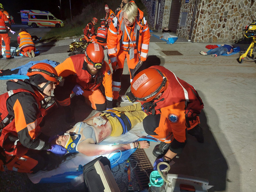 Ekipa štirih reševalcev oskrbuje ponesrečenko, ki leži na nosilih, ima vratno opornico in rano na trebuhu.