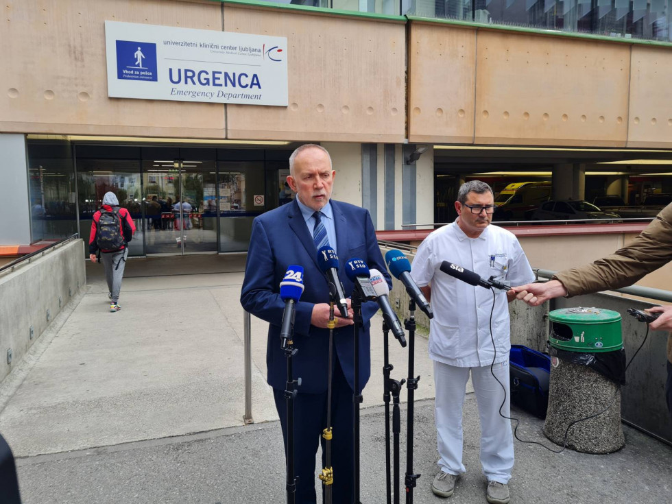 Dva moška, eden v temni obleki in drugi v zdravniški beli halji, stojita pred stavbo urgentnega centra. Pred njima so mikrofoni, zadaj se vidi napis Urgentnega centra.