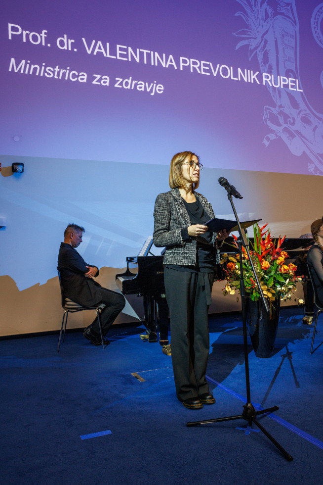 Ministrica stoji na odru pred mikrofonom, v rokah ima mapo, iz katere bere govor. v ozadju je klavir, za njim sedi pianist. V ozadju je vidna velika vaza z rožami.