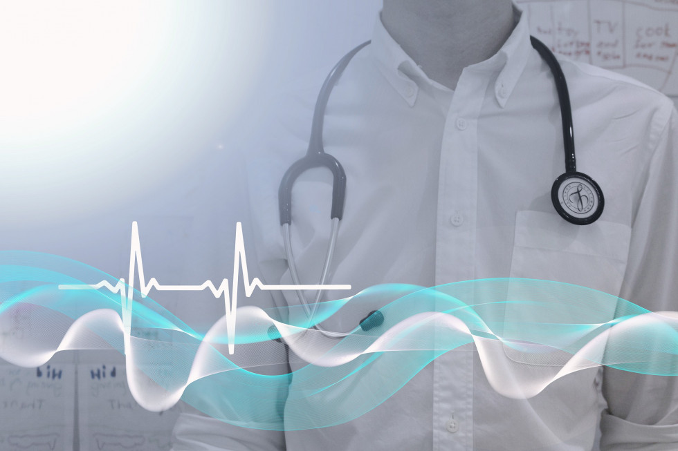 V ozadju fotografije je viden trup zdravnika v beli zdravniški halji s stetoskopom za vratom. V ospredju pa je grafika ritma srčnega utripa.