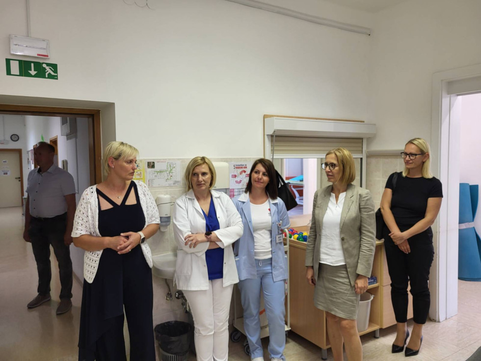 V ambulanti stoji pet žensk, dve sta v uniformi. Za njimi se vidi sprejemno okence. V ozadju je miza, na njej pisane kroglice. Na steni je umivalnik, pod njim pa koš.