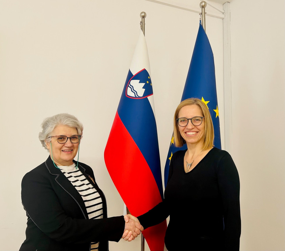 Dve ženski z očali se rokujeta pred slovensko zastavo in zastavo Evropske unije. Leva ima sive kratke lase, oblečena je v črn jopič in belo-črno črtasto majico. Desna ima do ramen segajoče svetle lase, oblečena je v črno majico. Obe sta nasmejani.