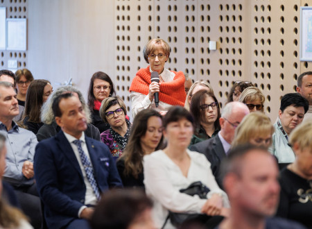 V sedeči publiki stoji ženska, ki v rokah drži mikrofon in vanj govori