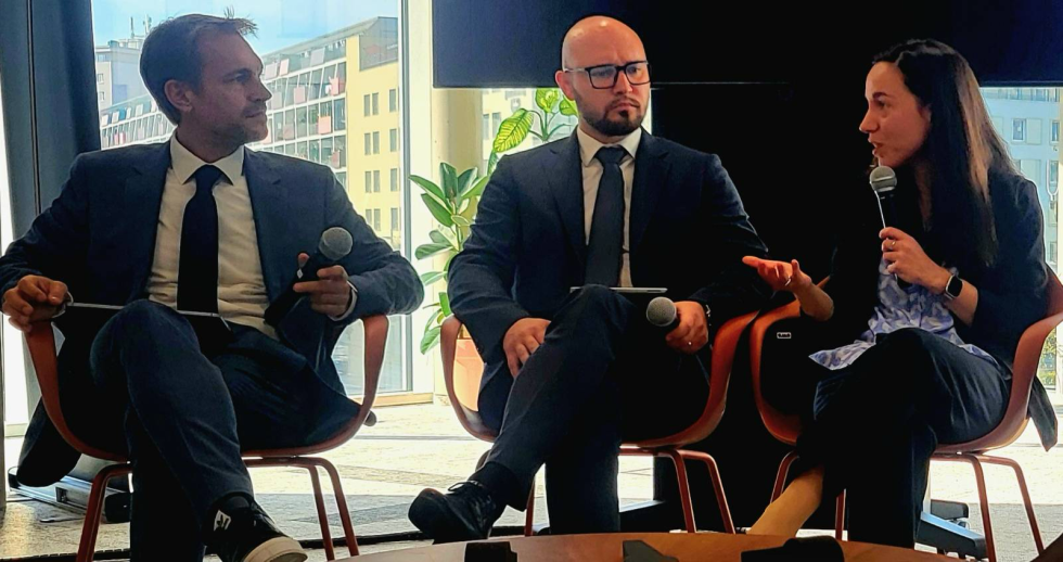 Državni sekretar Denis Kordež sedi za mizo med moderatorko in drugim govorcem
