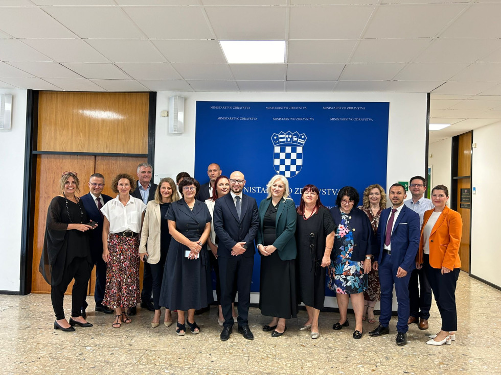 Pred steno s plakatom hrvaškega ministrstva za zdravje pozira skupina moških in žensk, vsi so nasmejani. V sredini sta slovenski in hrvaška državna sekretarja.