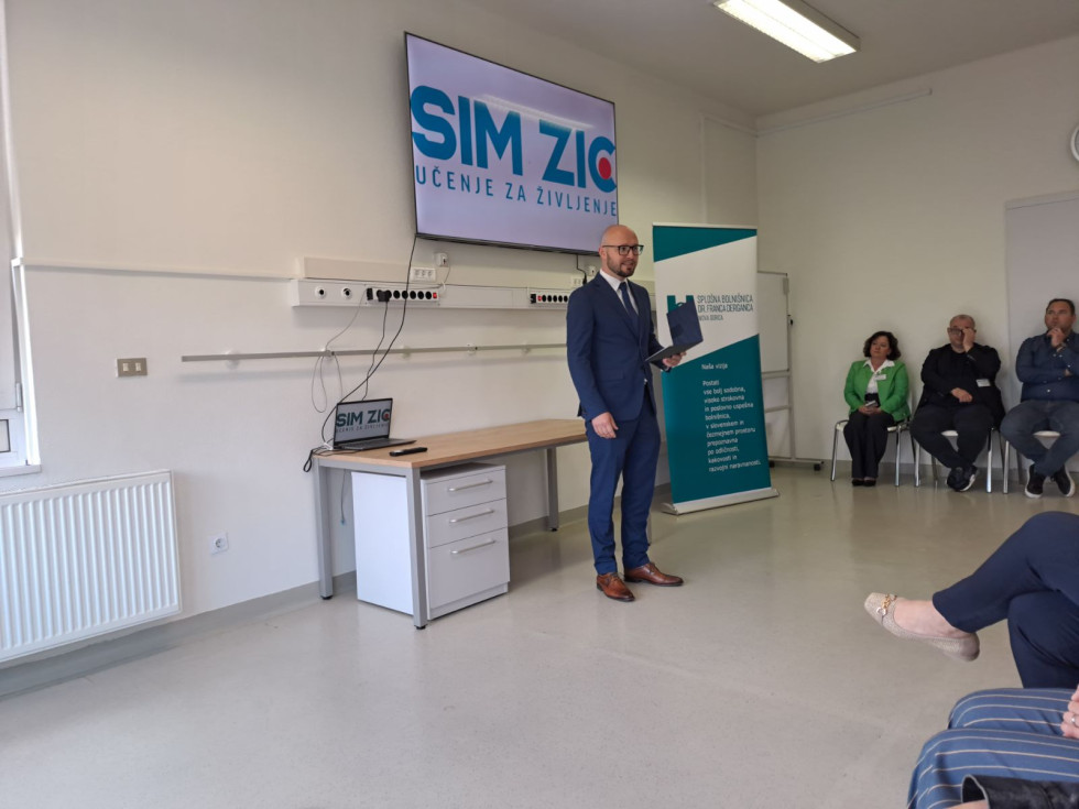 V sobi stoji moški v modri obleki z mapo v roki in govori. Za njim je miza z računalnikom, na steni pa monitor. Okoli njega sedijo ljudje in poslušajo.