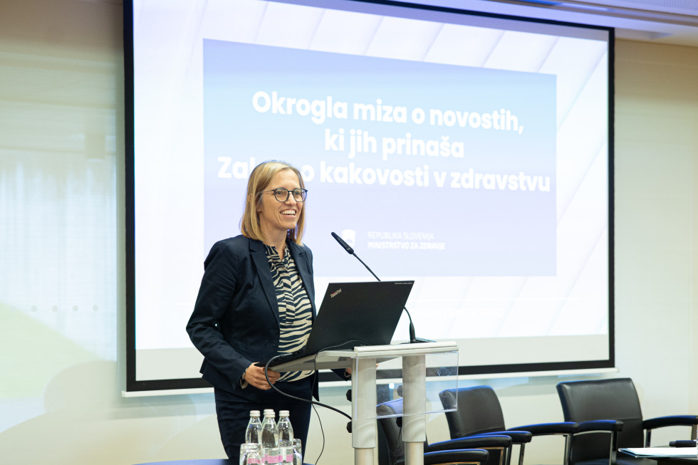 Za govorniškim pultom stoji ministrica in govori. Na pultu sta računalnik in mikrofon. V ozadju je projekcijsko platno, ob strani pa stoli. Ministrica je oblečena v moder suknjič in ima majico z zebrastim vzorcem. Nosi očala.