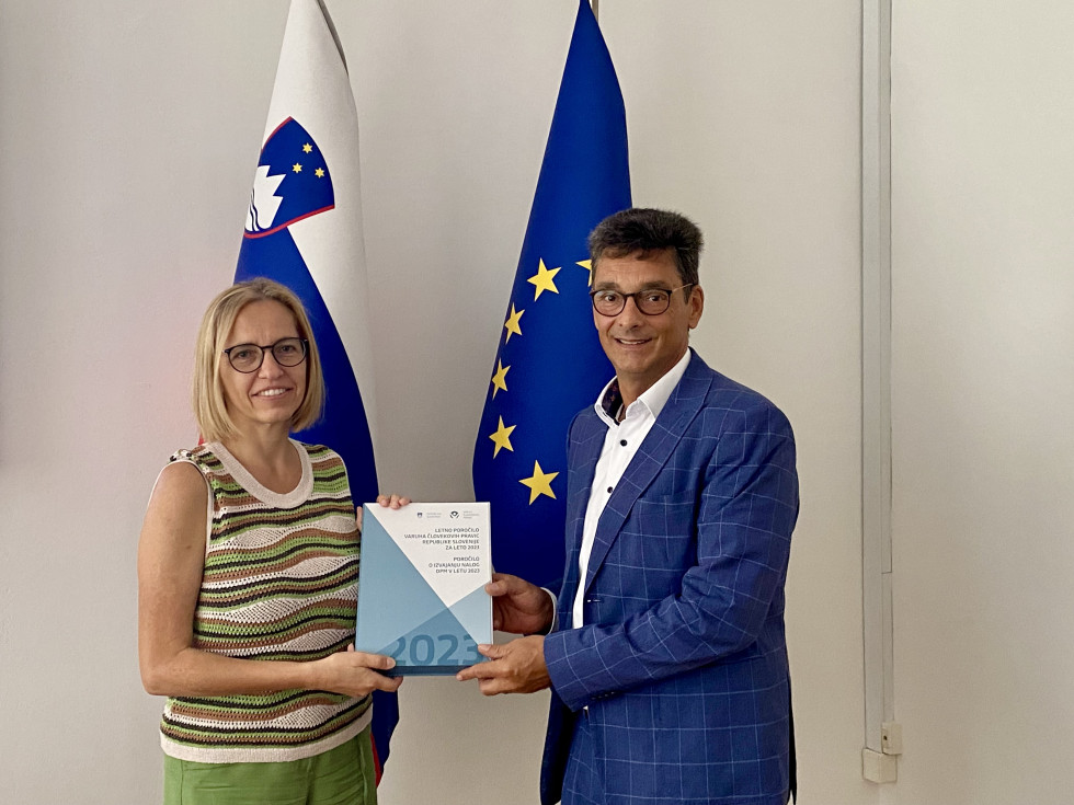 Ministrica in varuh pozirata pred slovensko zastavo in zastavo Evropske unije. Skupaj držita Poročilo. M9inistrica je oblečena v pleteno pisano majico brez rokavov in zelene hlače, Varuh pa v moder suknjič in belo srajco.