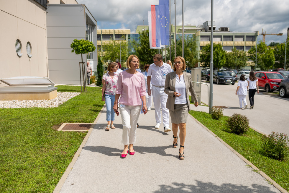 Ministrica in direktorica zdravstvenega doma hodita po pločniku, za njima je skupinica ljudi. Na desni inj zadaj so vidne sive in rumene stavbe bolnišnice, desno pa je parkirišče. Ob pločniku so tudi drogovi z zastavami.