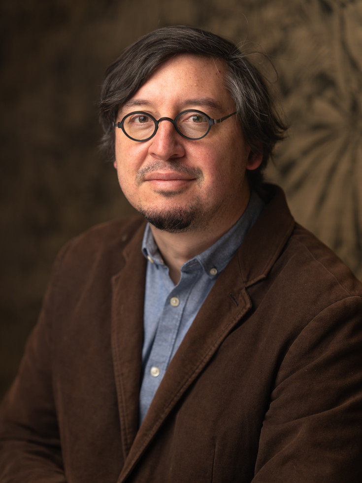 Portretna fotografija moškega srednjih let z očali. Oblečen je v rjav suknjič in modro srajco. Ima do ušes dolge temne lase in pristriženo bradico.