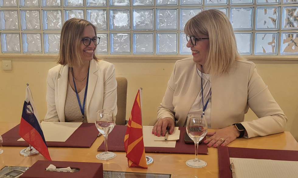 Za mizo sedita dve nasmejani ženski in se gledata. Na mizi pred njima so rdeče mape s papirji, slovenska in makedonska zastava, dva kozarca in mobilni telefon. Za njima je steklena stena.