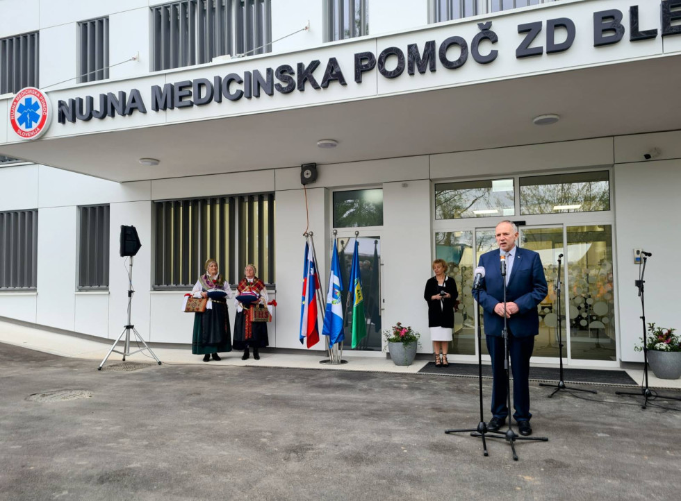 Pred stavbo z napisom Nujna medicinska pomoč državni sekretar govori v mikrofon. Za njim so vrata v stavbo, ob njih pa stoji ženska. Desno ob stavbi sta dve ženski v narodni noši, v rokah imata blazini. Zraven so postavljene zastave.