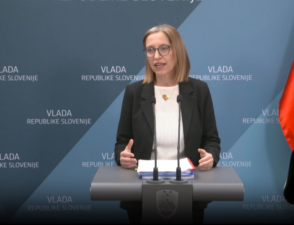 Ministrica za zdravje govori za govorniškim odrom v mikrofon. Oblečena je v črn suknjič in belo majico, nosi očala in zlato ogrlico, ima do ramen segajoče svetlo rjave lase. Za njo je stena, na kateri piše Vlada Republike Slovenije.