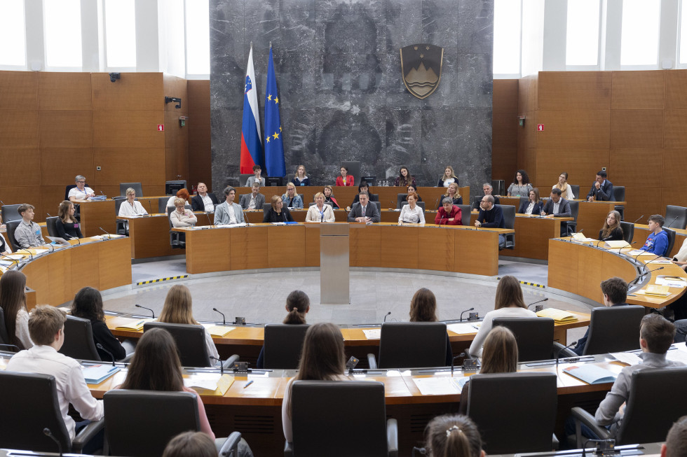 Dvorana z več vrstami stolov in miz v krogih. Na stolih sedijo ljudje in poslušajo moškega na sredini centralnega omizja, ki govori v mikrofon. Na sivi steni v ozadju je grb Republike Slovenije. Zraven sta tudi zastava Evropske unije in slovenska zastava.