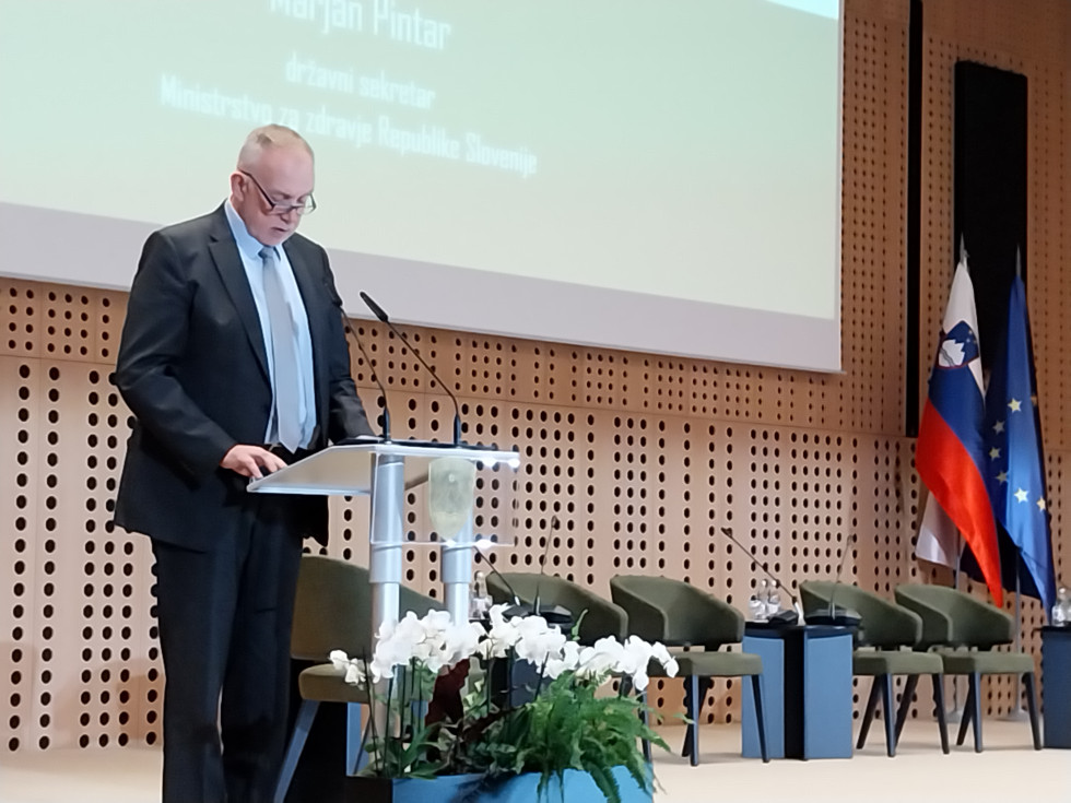 Moški v sivi obleki stoji za govorniškim pultom na odru. Pred pultom je cvetlični aranžma. Na odru so prazni stoli, v kotu pa slovenska zastava in zastava Evropske unije.