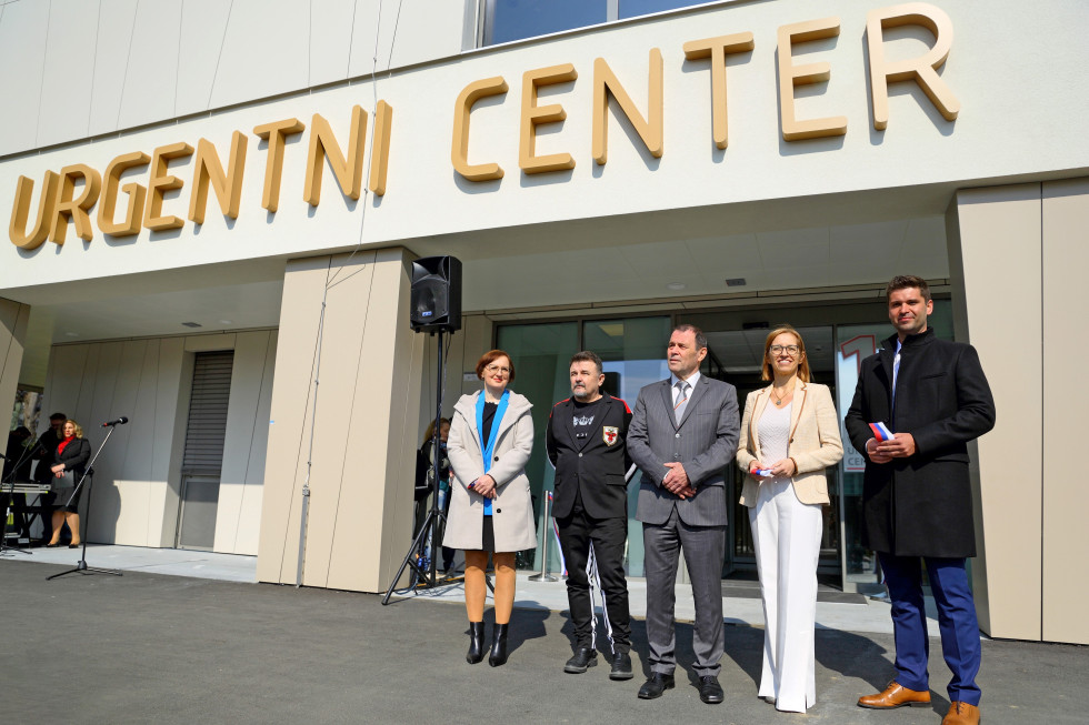 Pred stavbo, na kateri z velikimi črkami piše urgentni center, stojijo dve ženski in trije moški. Za njimi je viden zvočnik, levo od njih pa mikrofon.