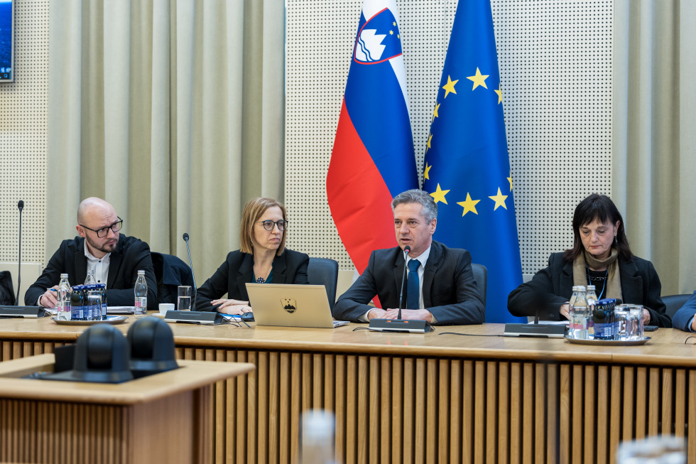 Za leseno mizo sedijo dva moška in dve ženski. Za njimi sta slovenska zastava in zastava Evropske unije. Eden od moških govori v mikrofon, ostali trije ga gledajo in poslušajo. Na mizi je tudi siv računalnik s slovenskim grbom.