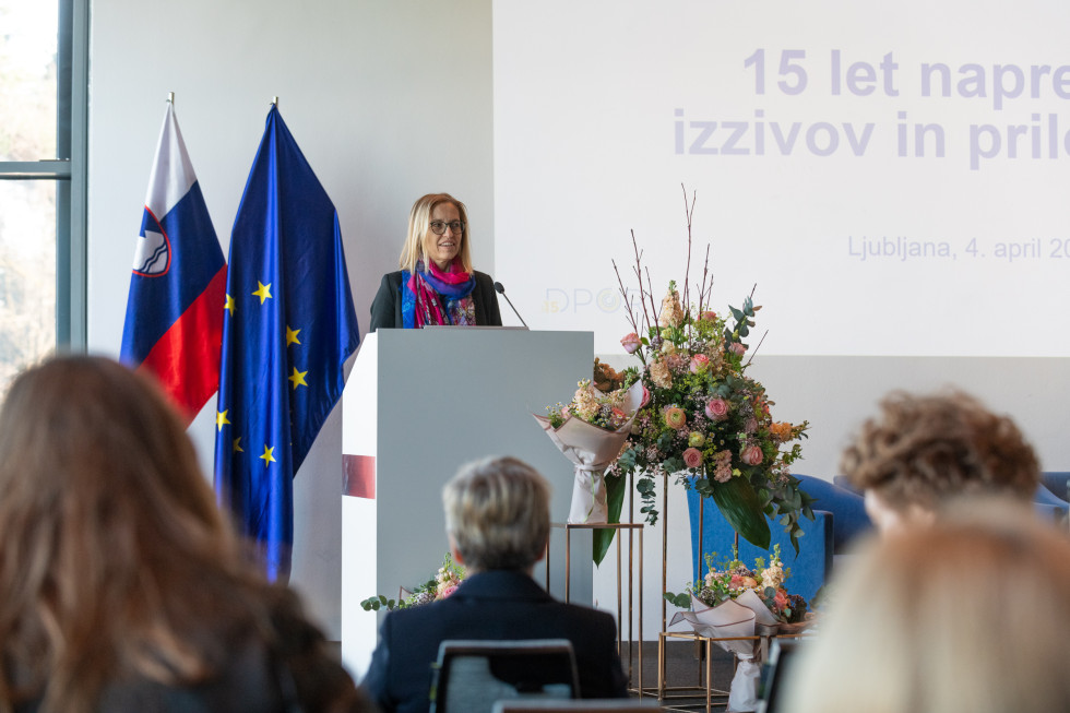 Ministrica govori v mikrofon za govorniškim pultom. Pred njo je na stojalih več večjih šopkov pisanih rož. V ospredju je nekaj zabrisanih glav udeležencev dogodka. Za ministrico sta evropska in slovenska zastava.