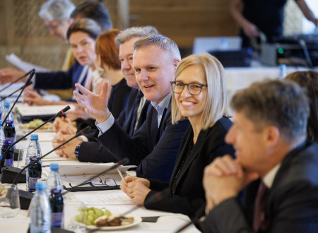 Ministrica v temnem ženskem kostimu sedi za dolgo mizo, ob njej do drugi predstavniki vlade