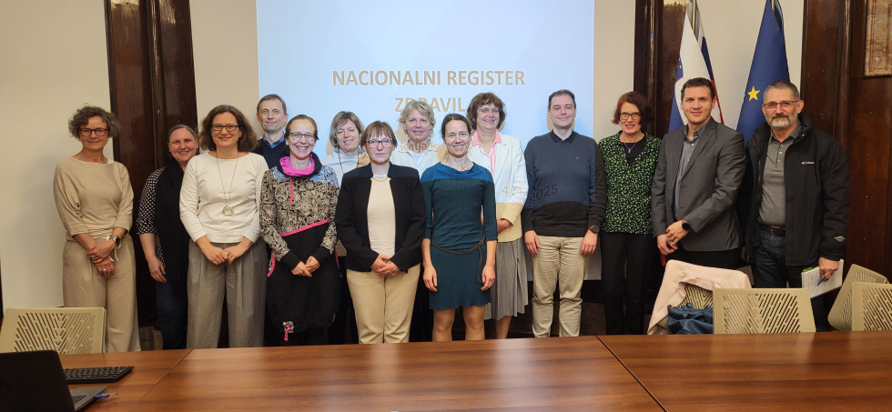 Skupina štirinajstih ljudi pozira pred projekcijskim platnom, na katerem piše Nacionalni register zdravil. Pred njimi so dolga lesena miza in bež plastični stoli.