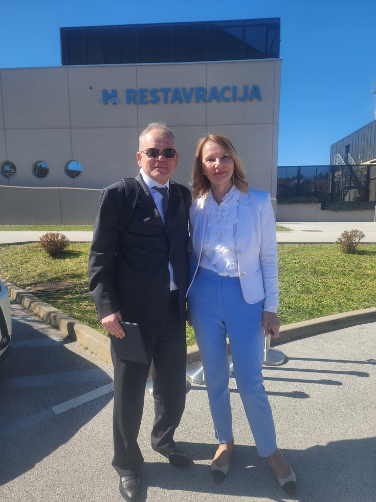 Državni sekretar in direktorica bolnišnice v jasnem, sončnem dnevu na parkirišču pozirata pred betonsko stavbo, na kateri sta napis restavracija in logotip bolnišnice.