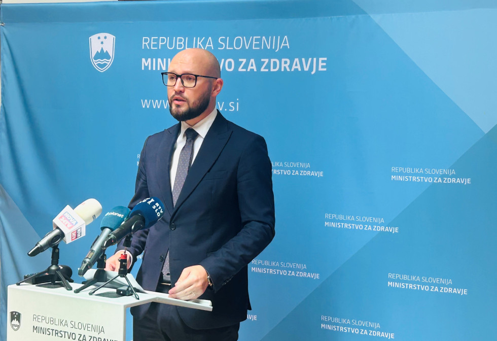 Državni sekretar stoji za govorniškim pultom, na katerem so mikrofoni. Oblečen je v modro obleko, belo srajco in temno kravato z belimi pikami. Nosi očala. Za njim je modra stena z logotipi ministrstva za zdravje.
