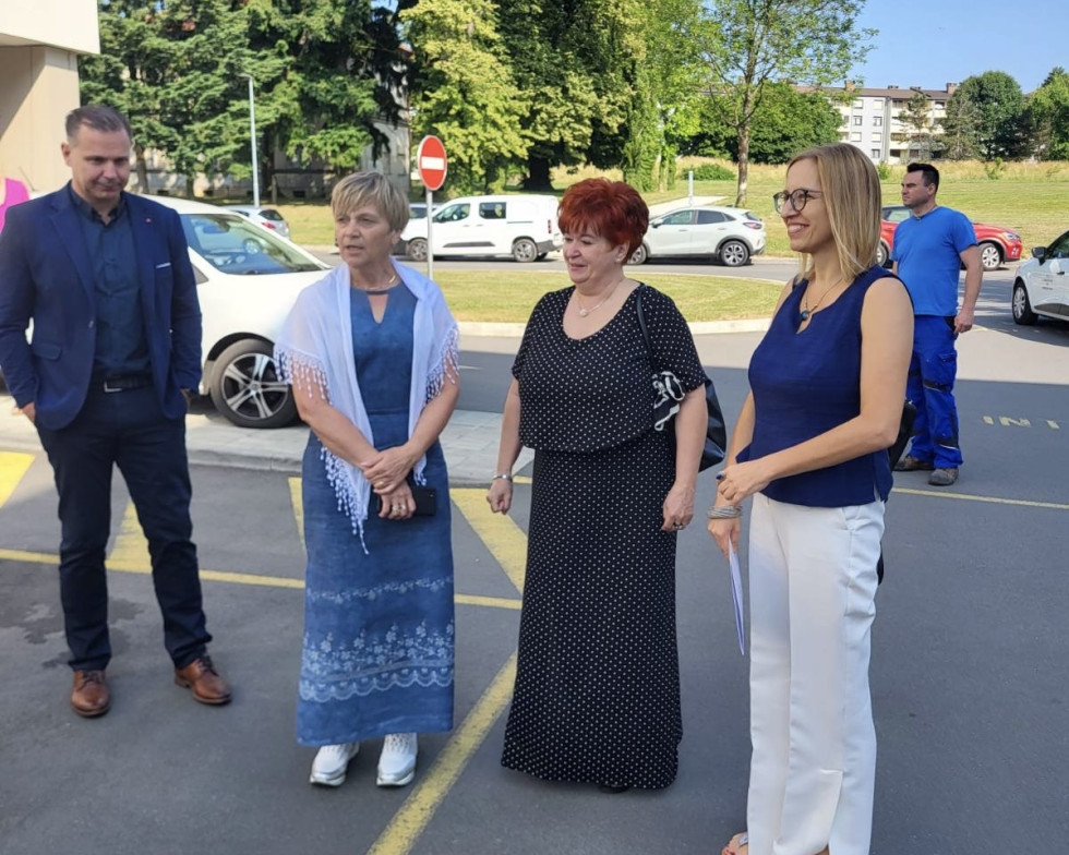 Zunaj na parkirišču stojijo tri ženske, v ozadju sta še dva moška. Je lep sončen dan, ženske so nasmejane in slavnostno oblečene. Za njimi so parkirani avtomobili, v ozadju je park.