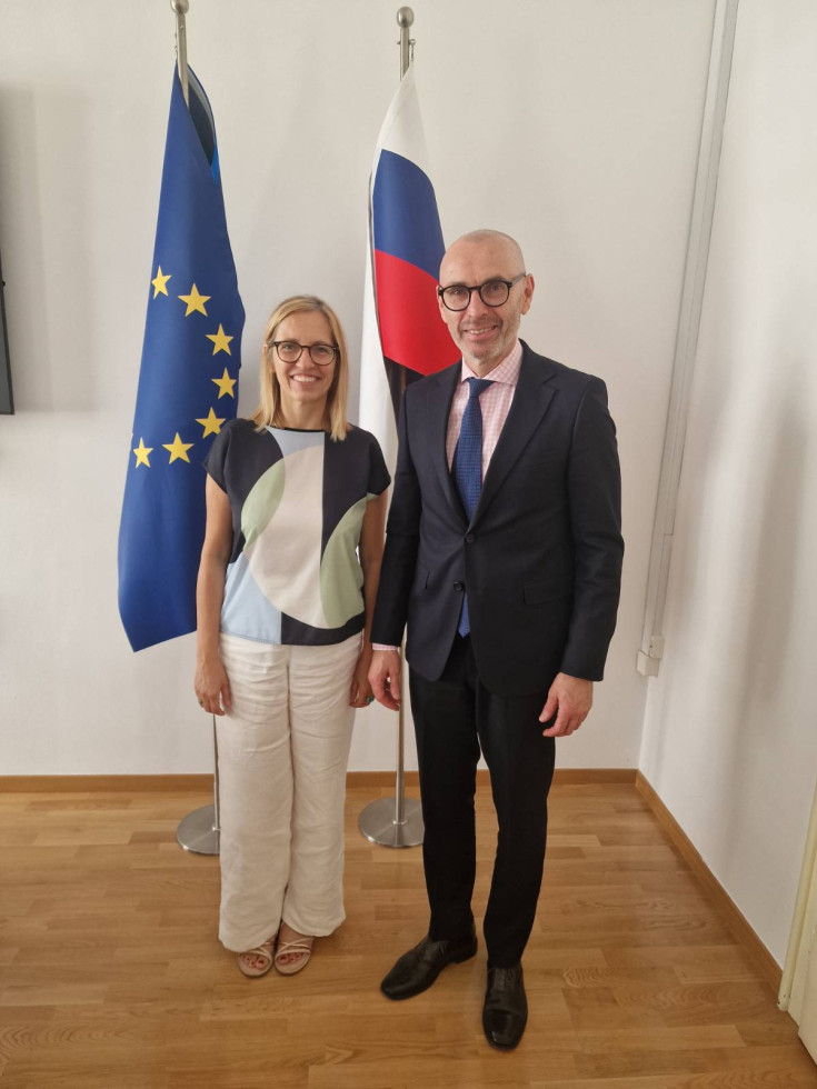 Ministrica in predstavnik SZO nasmejana pozirata pred zastavama Evropske unije in republike Slovenije. Ministrica nosi pisano majico in bele hlače, predstavnik pa črno obleko, rdečo karirasto srajco in modro kravato.