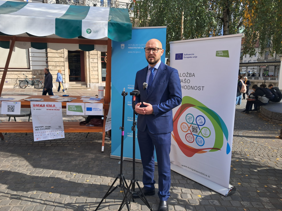 Državni sekretar zunaj na tlakovanem trgu govori v mikrofona. Za njim so stojnica in dva velika plakata. Zadaj je videti več ljudi.