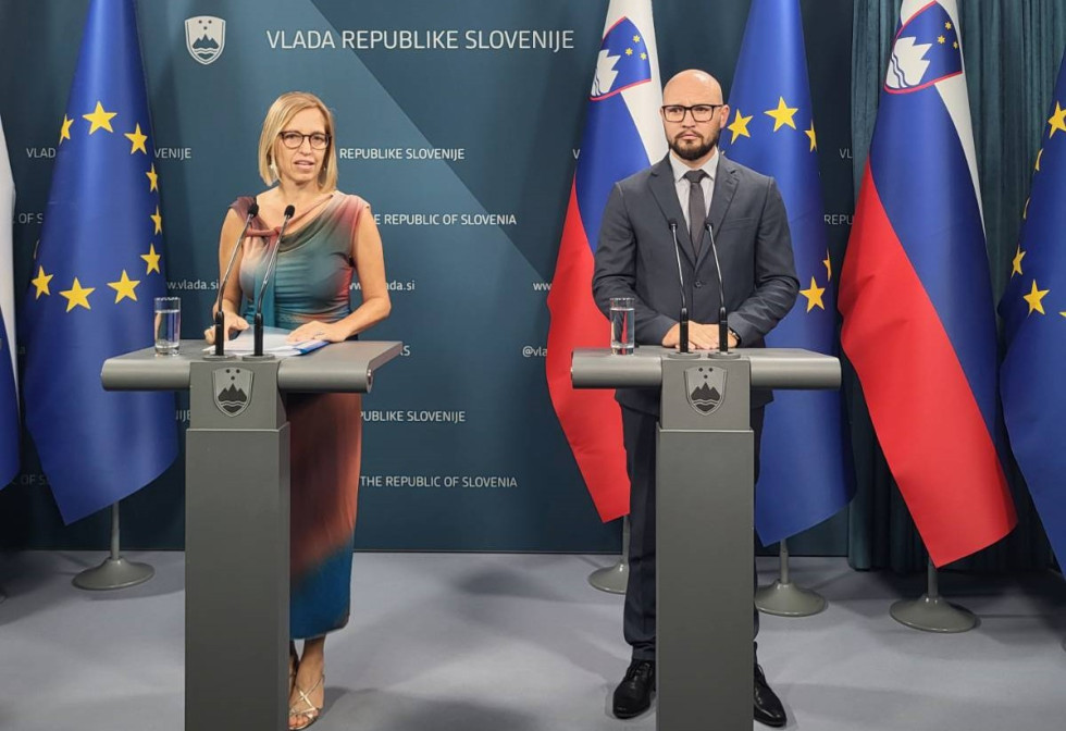Ministrica in državni sekretar stojita za govorniškima odroma. Ministrica govori v mikrofon. Za njima so slovenske zastave in zastave Evropske unije ter stena z grbom Vlade Republike Slovenije.