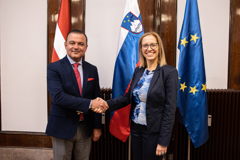 Ministra se rokujeta pred slovensko in latvijsko zastavo ter zastavo Evropske unije.